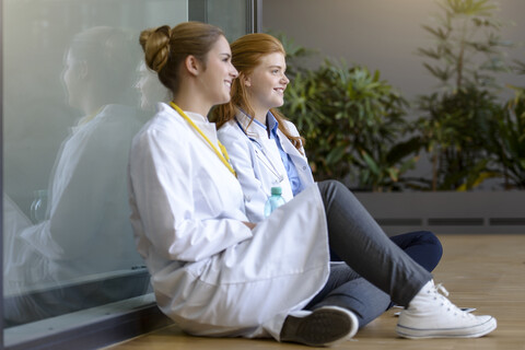 Zwei junge Ärztinnen sitzen auf dem Boden am Krankenhauseingang, lizenzfreies Stockfoto