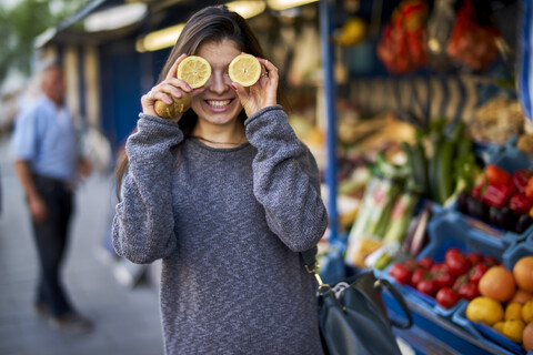 Lachende junge Frau auf dem Markt, die ihre Augen mit Zitronenhälften bedeckt, lizenzfreies Stockfoto