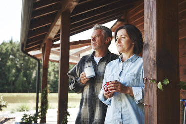 Mature couple standing on cabin veranda, holding tin cups, looking at view - CUF10653