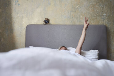 Frau im Bett, versteckt unter der Decke, Arm in der Luft, Hand mit Friedenszeichen - CUF10672