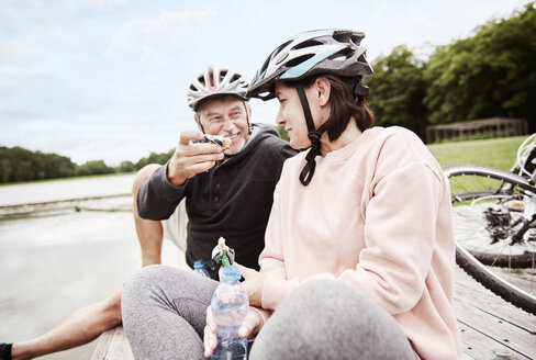 Mature couple relaxing on jetty, enjoying snack, bicycles behind them - CUF10688