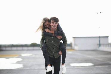 Young man giving girlfriend a piggyback across car park - CUF10731