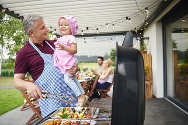 Mature man carrying toddler granddaughter barbecuing at family lunch on patio - CUF10869