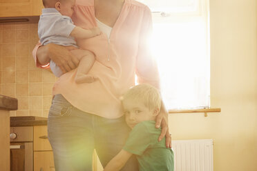 Junge umarmt das Bein der Mutter, während sie seinen kleinen Bruder in der Küche trägt - CUF10884
