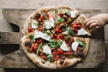 Sliced pizza on chopping board, child's hand reaching for pizza slice, overhead view - CUF11119