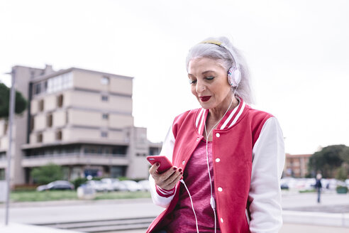 Mature woman in baseball jacket listening to headphones and looking at smartphone - CUF11284