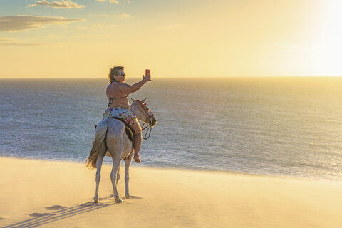Frau reitet auf einem Pferd am Strand und fotografiert die Aussicht mit ihrem Smartphone, Jericoacoara, Ceara, Brasilien, Südamerika, lizenzfreies Stockfoto