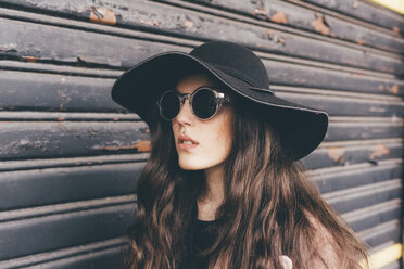 Portrait of young woman standing beside shutter, wearing floppy hat and sunglasses - CUF11413