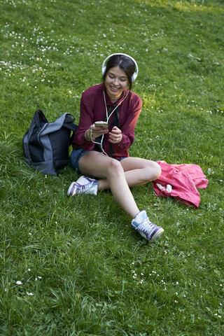 Junge Frau mit Kopfhörern, die auf einer Wiese sitzt und ein Mobiltelefon benutzt, lizenzfreies Stockfoto