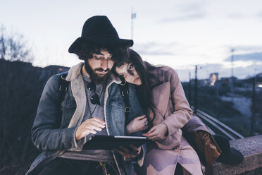 Junges Paar sitzt in der Abenddämmerung an einer Wand und schaut auf ein digitales Tablet - CUF11437