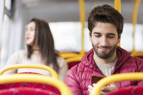 UK, London, Porträt eines lächelnden jungen Mannes, der im Bus ein Handy benutzt, lizenzfreies Stockfoto