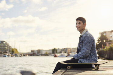 Junger Mann sitzt am Fluss, Skateboard neben ihm, Bristol, UK - CUF11881