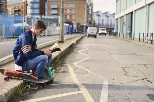Junger Mann sitzt auf dem Bordstein und liest ein Buch, neben ihm ein Skateboard, Bristol, UK - CUF11884