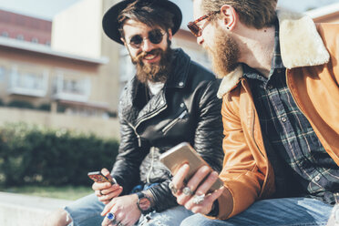 Zwei junge männliche Hipster sitzen mit Smartphones an der Wand - CUF12031