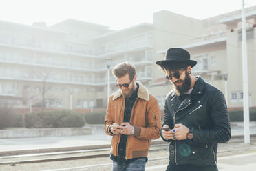 Zwei junge männliche Hipster schauen an der Straßenbahnhaltestelle auf ihre Smartphones - CUF12032