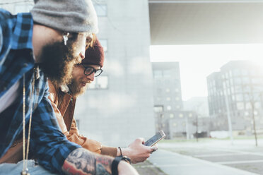 Zwei junge männliche Hipster sitzen an der Wand und schauen auf ihr Smartphone, Seitenansicht - CUF12037