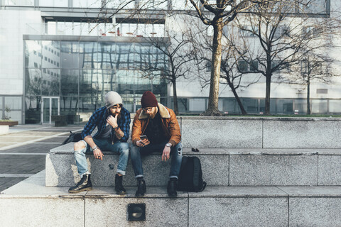 Zwei junge männliche Hipster sitzen an der Wand und schauen auf ihr Smartphone, lizenzfreies Stockfoto