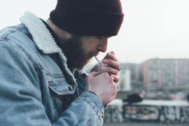 Young male hipster in knit hat igniting cigarette in city - CUF12049