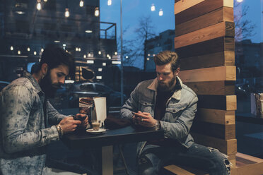 Zwei junge männliche Hipster in einem Straßencafé, die nachts auf ihr Smartphone schauen - CUF12052