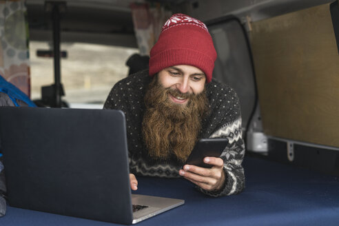 Portrait of smiling hipster lying in van using smartphone and laptop - AFVF00506