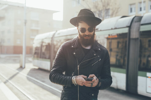 Junger männlicher Hipster, der an einer Straßenbahnhaltestelle auf sein Smartphone schaut - CUF12191