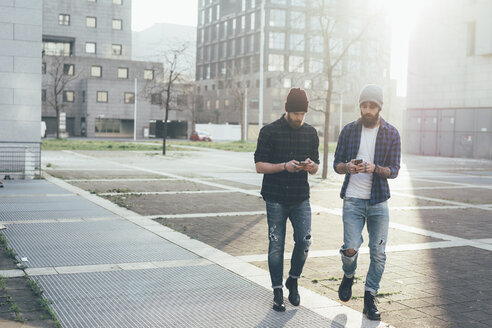 Zwei junge männliche Hipster, die in der Stadt spazieren gehen und dabei auf ihre Smartphones schauen - CUF12195