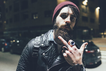 Porträt eines jungen männlichen Hipsters auf einer nächtlichen Straße in der Stadt, der eine obszöne Fingergeste macht - CUF12200