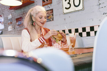 Ältere Frau in Baseballjacke isst Burger in einem Diner der 1950er Jahre - CUF12358
