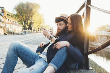 Hipster-Paar sitzt am Stadtkanal und schaut auf sein Smartphone - CUF12424
