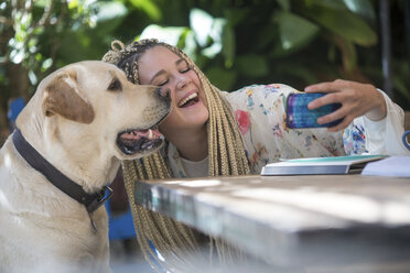 Junge Frau mit Dreadlocks, die im Freien sitzt und ein Selfie mit ihrem Hund macht und ein Smartphone benutzt - CUF12475