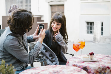 Junges Paar sitzt vor einem Café, Mann fotografiert Frau mit Smartphone - CUF12564