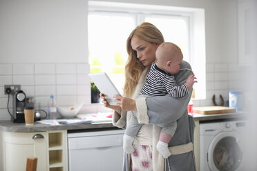 Mutter steht in der Küche, hält einen kleinen Jungen und schaut auf ein digitales Tablet - CUF12569
