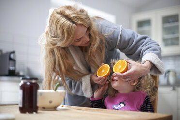 Junges Mädchen sitzt am Küchentisch, Mutter hält halbierte Orange vor die Augen der Tochter - CUF12572