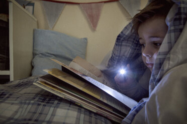 Boy lying in bed wrapped in duvet reading book by torchlight - ISF02107