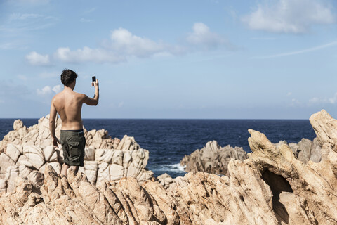 Rückansicht eines jungen Mannes auf einem Felsen, der mit seinem Smartphone den Horizont über dem Meer fotografiert, Costa Paradiso, Sardinien, Italien, lizenzfreies Stockfoto