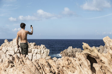 Rückansicht eines jungen Mannes auf einem Felsen, der mit seinem Smartphone den Horizont über dem Meer fotografiert, Costa Paradiso, Sardinien, Italien - CUF13018