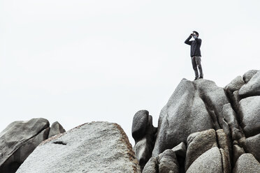 Low angle view of young man standing on rocks, pretending to use binoculars, Costa Smeralda, Sardinia, Italy - CUF13025