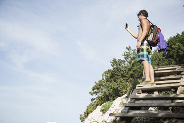 Niedrige Seitenansicht eines jungen Mannes, der auf einer Holztreppe steht und ein Smartphone zum Fotografieren benutzt, Cala Luna, Sardinien, Italien - CUF13041