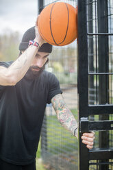 Mittlerer erwachsener Mann, der am Zaun steht und einen Basketball gegen den Zaun hält, nachdenklicher Ausdruck - CUF13086