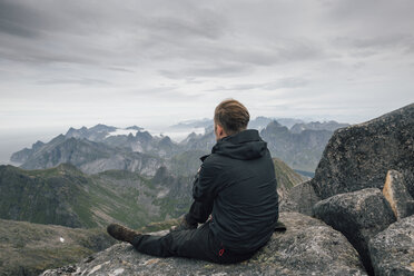 Norwegen, Lofoten, Moskenesoy, Junger Mann sitzt am Hermannsdalstinden und blickt über den Kjerkefjord - GUSF00828