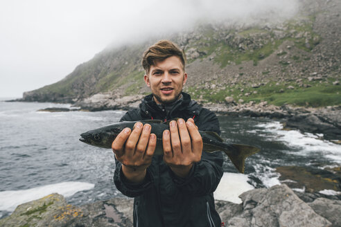 Norwegen, Lofoten, Moskenesoy, Junger Mann hält fangfrischen Fisch am Strand von Horseid - GUSF00853