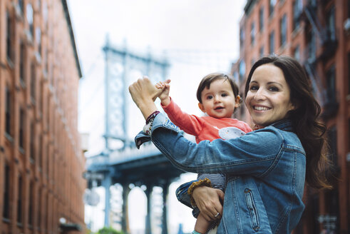 USA, New York, New York City, Glückliche Mutter hält ein Baby in Brooklyn mit der Manhattan Bridge im Hintergrund - GEMF02005