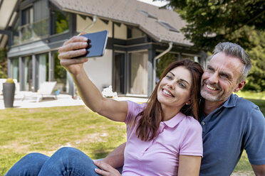 Happy couple sitting in garden of their home taking a selfie - DIGF04369