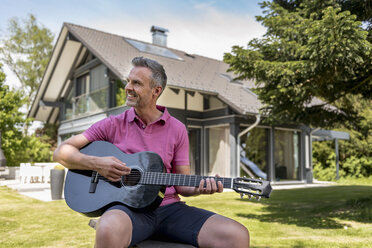 Smiling mature man sitting in garden of his home playing guitar - DIGF04371