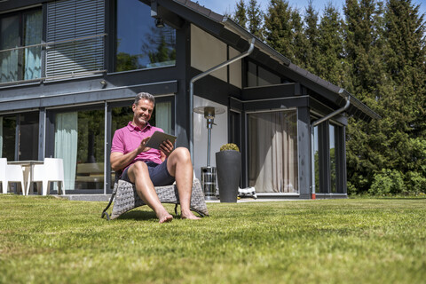 Lächelnder reifer Mann, der im Garten seines Hauses sitzt und ein Tablet benutzt, lizenzfreies Stockfoto