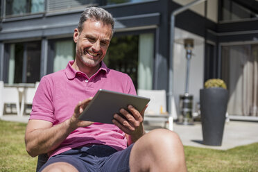 Lächelnder reifer Mann, der im Garten seines Hauses sitzt und ein Tablet benutzt - DIGF04375