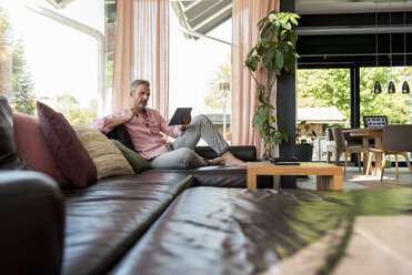 Mature man sitting on couch at home using a tablet - DIGF04383