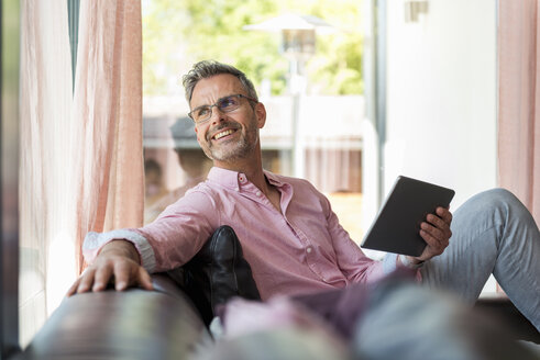 Lächelnder reifer Mann sitzt auf der Couch zu Hause und hält ein Tablet - DIGF04384
