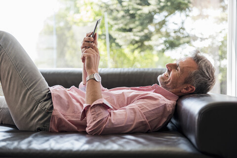 Lächelnder reifer Mann, der zu Hause auf der Couch liegt und ein Mobiltelefon benutzt, lizenzfreies Stockfoto