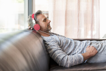 Relaxed mature man lying on couch at home wearing headphones - DIGF04388